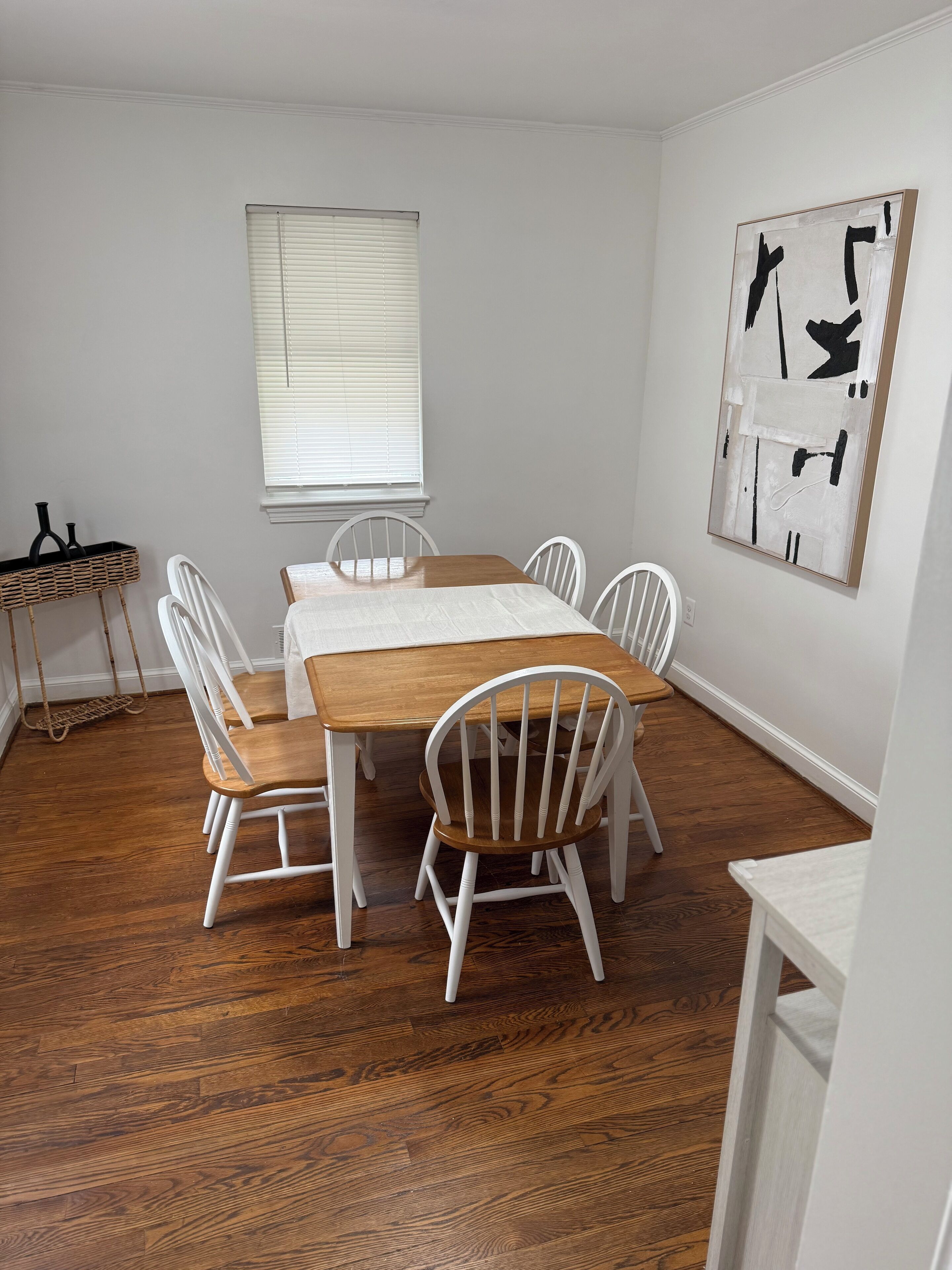 A dining area with a wooden table and white chairs, a window with blinds, and a wall-mounted abstract painting.