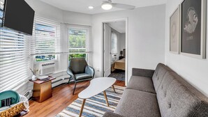 Un salon moderne avec un canapé gris, une table basse blanche, un fauteuil en cuir noir et un téléviseur fixé au mur. Les fenêtres sont équipées de stores blancs, il y a un ventilateur de plafond et une œuvre d’art encadrée est accrochée au mur.