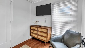 A room with a wooden dresser, a leather chair, a flat-screen TV mounted on the wall, and a window with blinds.