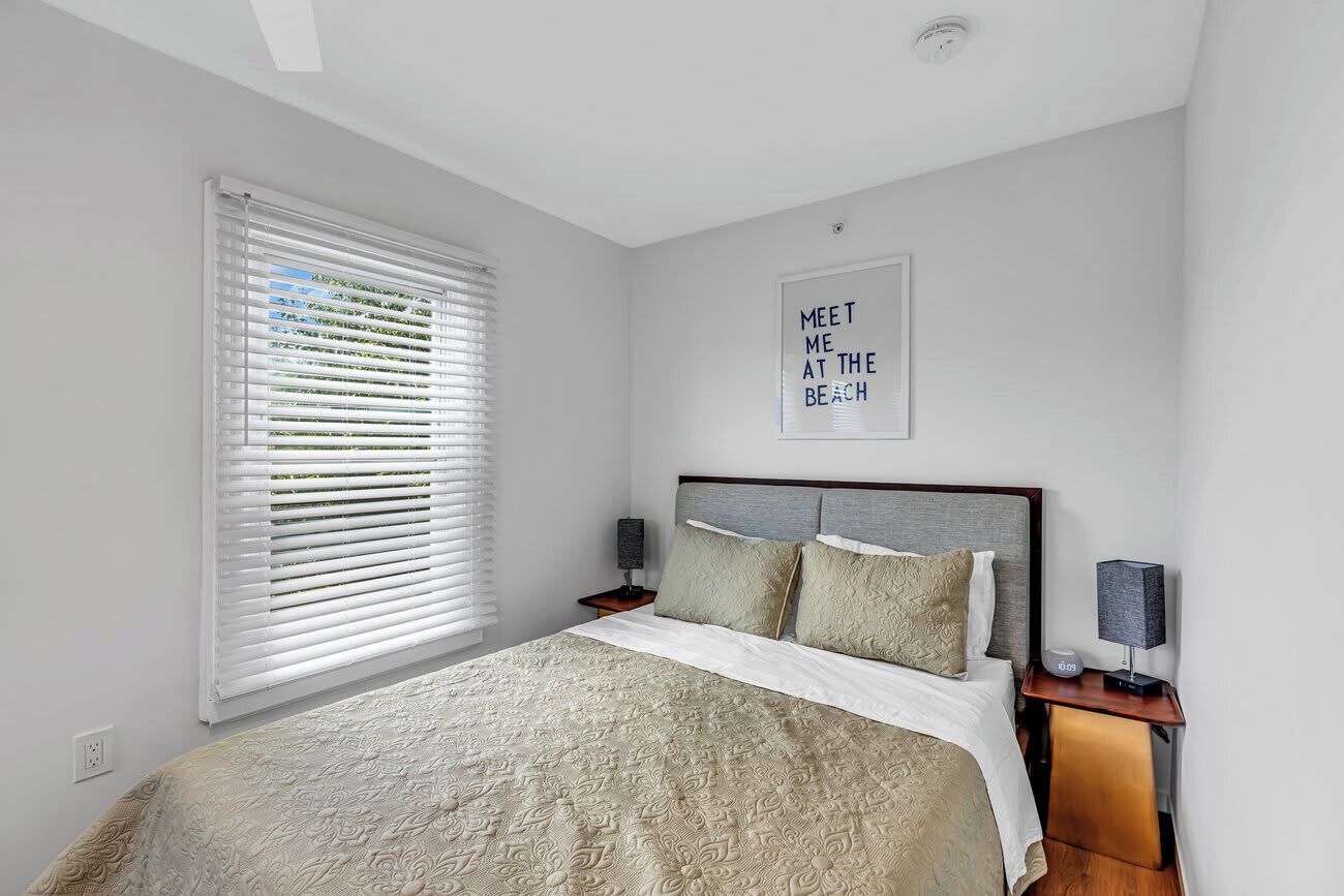 A modern bedroom with a large bed, a wall-mounted artwork, a bedside table with a lamp, and a window with blinds.