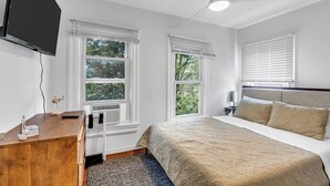 A bedroom with a bed, a desk, a TV, and windows with blinds.