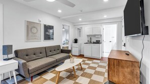 A modern living room with a sofa, a coffee table, and a TV mounted on the wall. There is a kitchen area with cabinets and a ceiling fan.