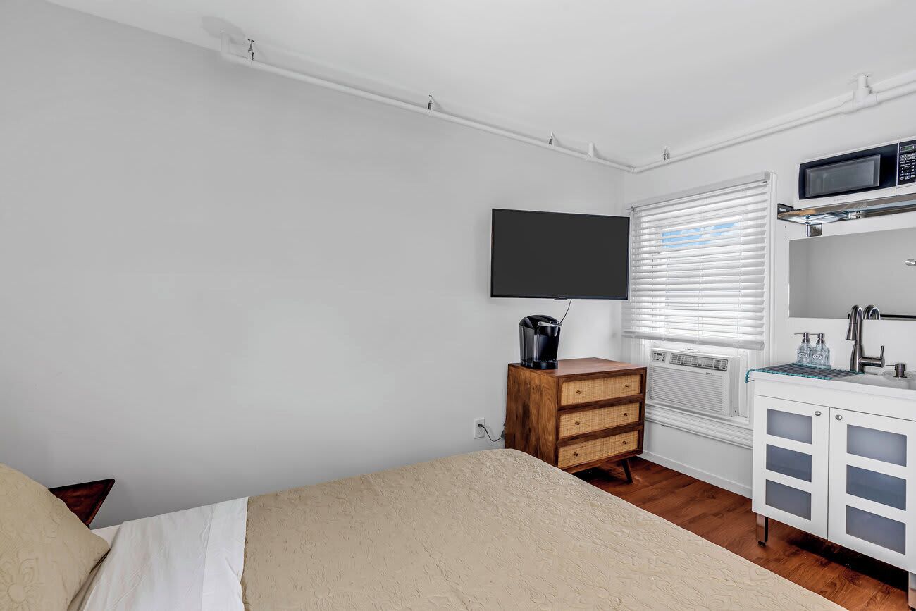 Ein Schlafzimmer mit einem Bett, einer Holzkommode, einem Fernseher, einer Mikrowelle und einem Fenster mit Jalousien.