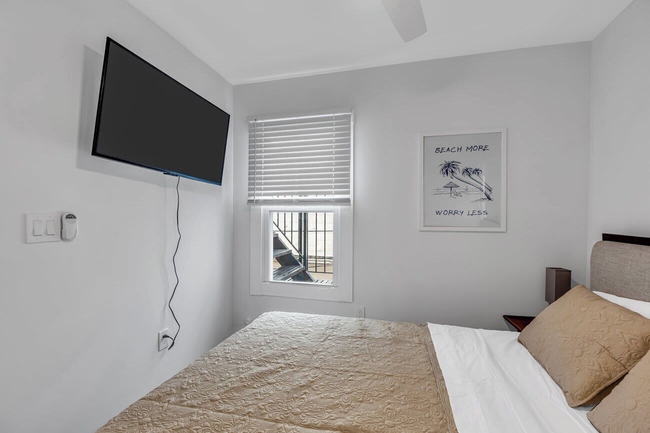 A bedroom with a bed, a TV mounted on the wall, a window with blinds, and a poster on the wall.