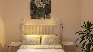 A bedroom with a bed, two bedside tables, a radiator, a wall-mounted abstract painting, and a potted plant.