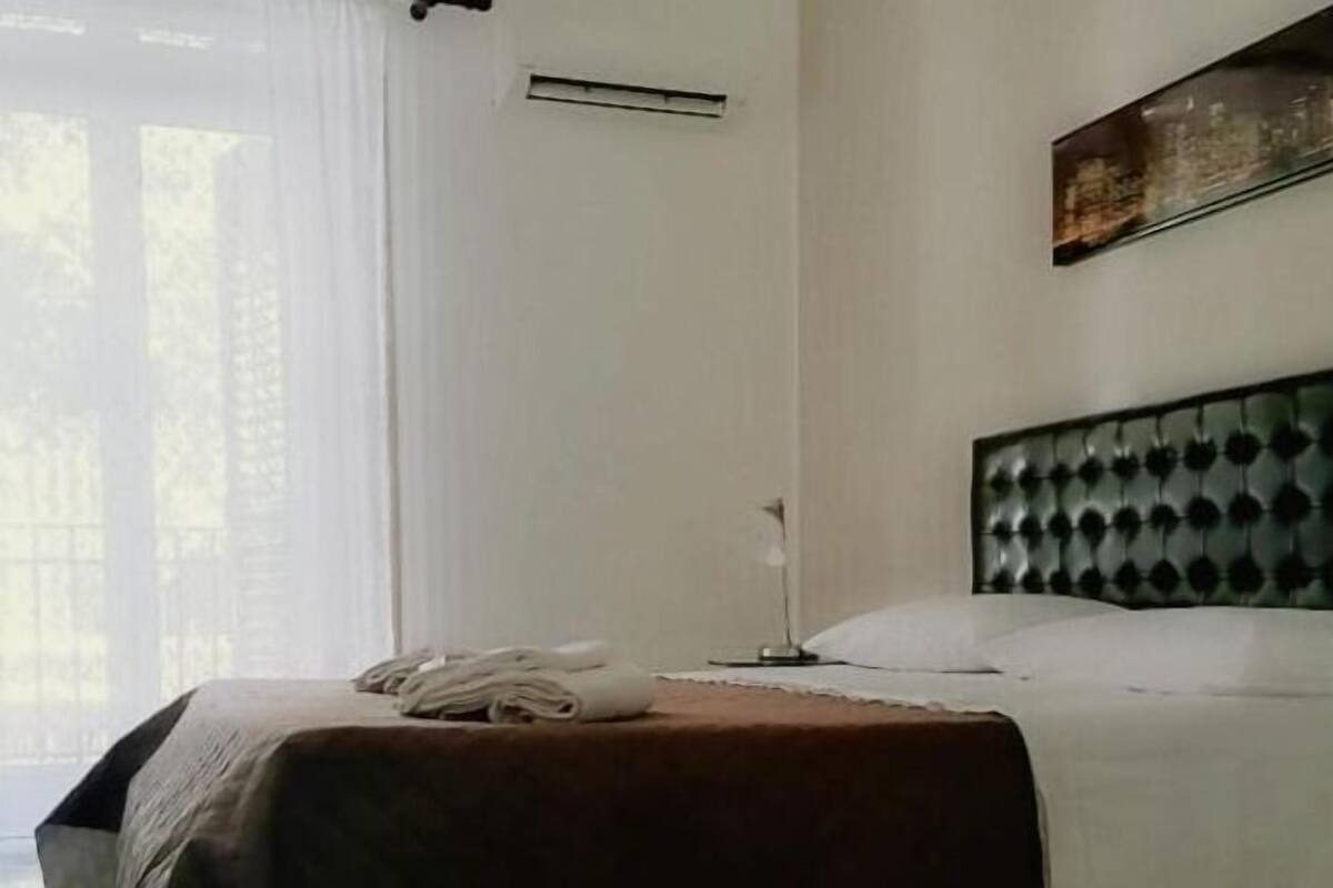 Una habitación de hotel con cama, mesita de noche, lámpara y ventana con cortinas.