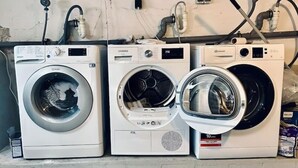 Three front-loading washing machines on a shelf in a utility room with visible plumbing and electrical connections.