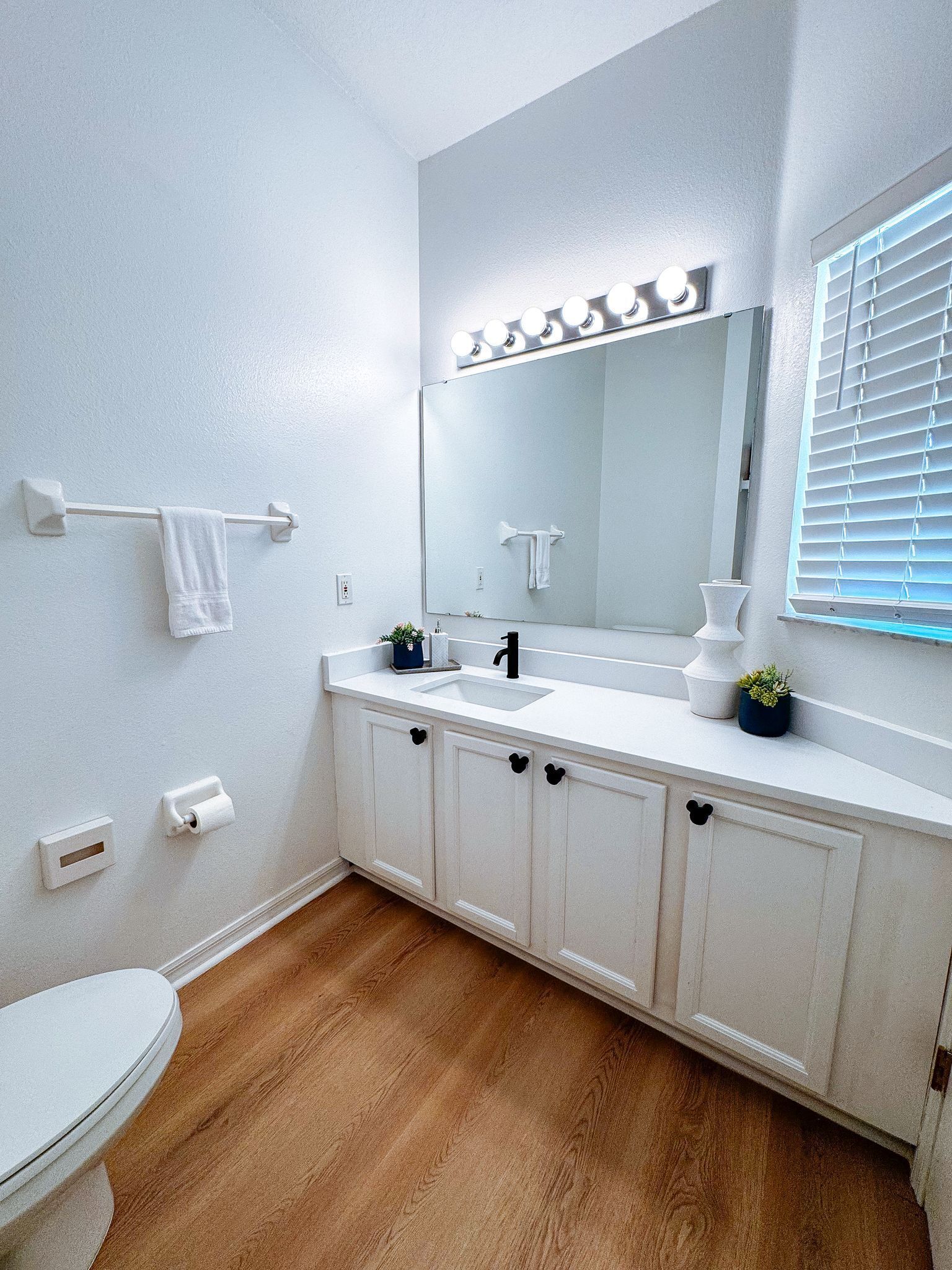 A bathroom with a white vanity, a large mirror with lights, a towel rack, and a toilet.