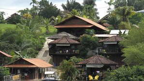 A multi-story wooden house with a boat docked in front, surrounded by lush greenery and palm trees.