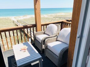 A wooden deck with wicker furniture and a table, overlooking a beach. - Sea Breeze Beautiful Oceanfront Townhome in Kure (Kure Beach)