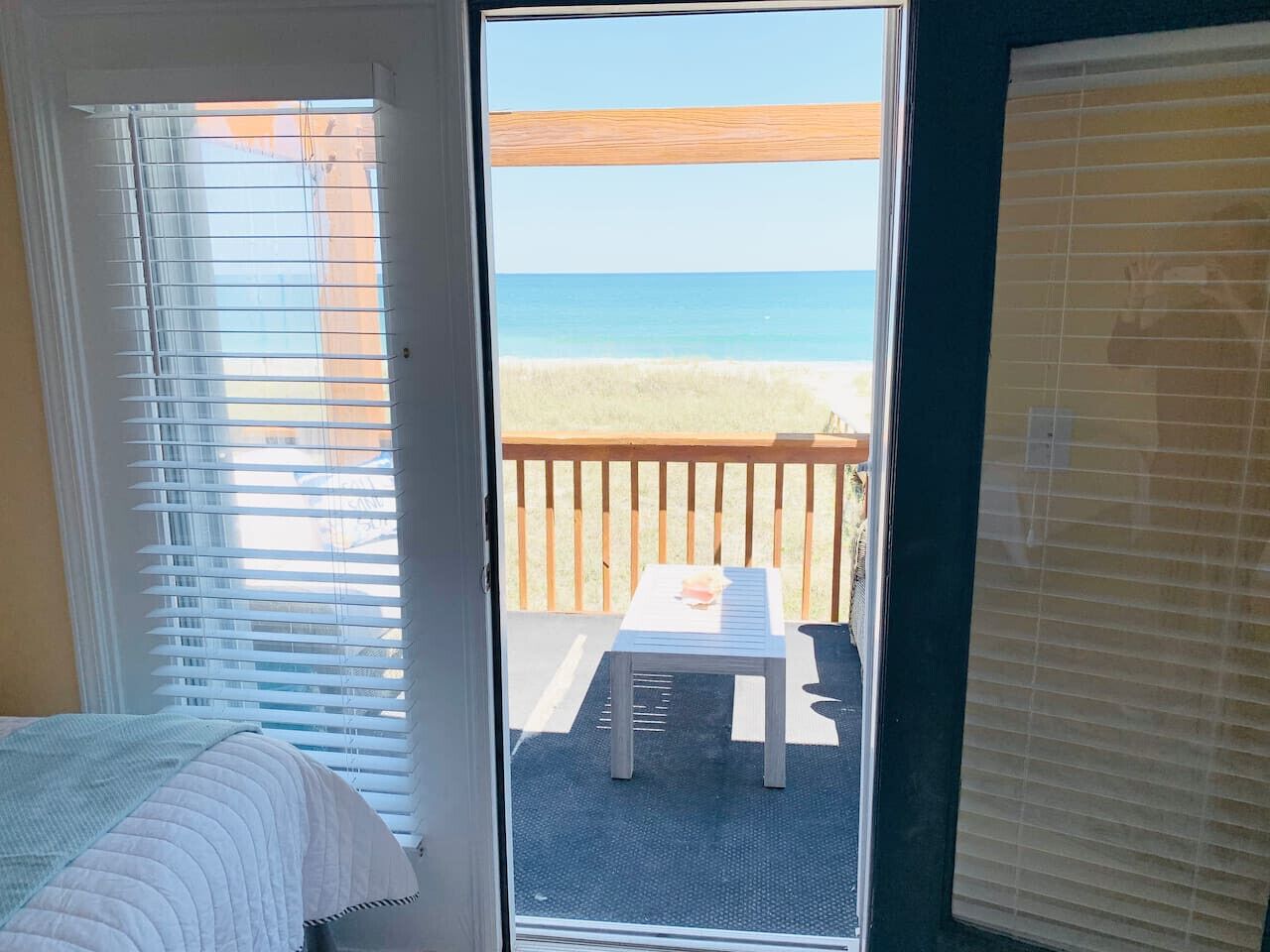 Una habitación con cama, ventana con persianas y vista a una playa con mesa y sillas afuera.