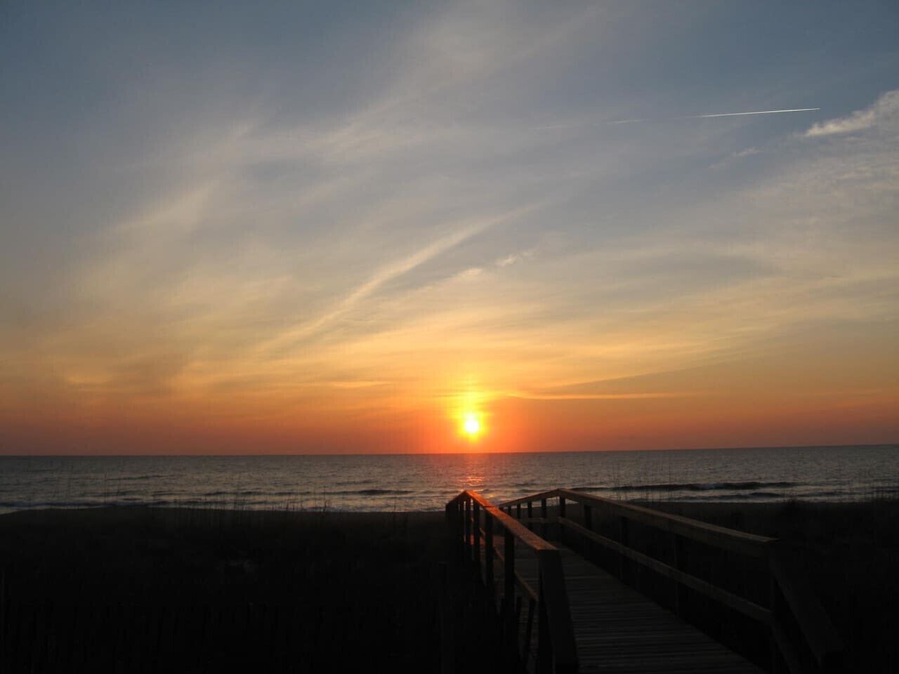 A sunset over the ocean with a wooden pier leading to the beach.