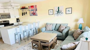 Een woonkamer met een bank, een salontafel en een eethoek met stoelen, met een keuken op de achtergrond.