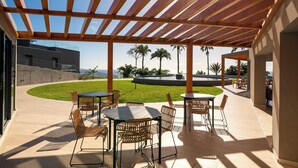 An outdoor patio area with wooden pergola, tables, and chairs, overlooking a landscaped garden and a body of water. - With pool in Funchal - Acqua Residence V (Funchal)