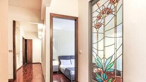 Un couloir au plancher de bois mène à une chambre à coucher où se trouvent un lit et une table de chevet. Une grande fenêtre à vitraux aux motifs floraux est visible sur le mur de droite.