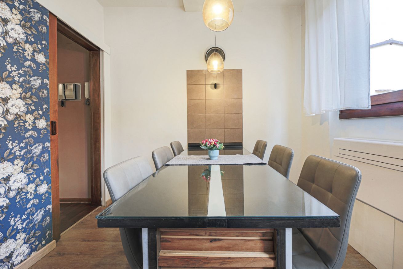 Un coin repas comprenant une table en verre, six chaises, un vase avec des fleurs, un luminaire fixé au mur et une porte en bois.