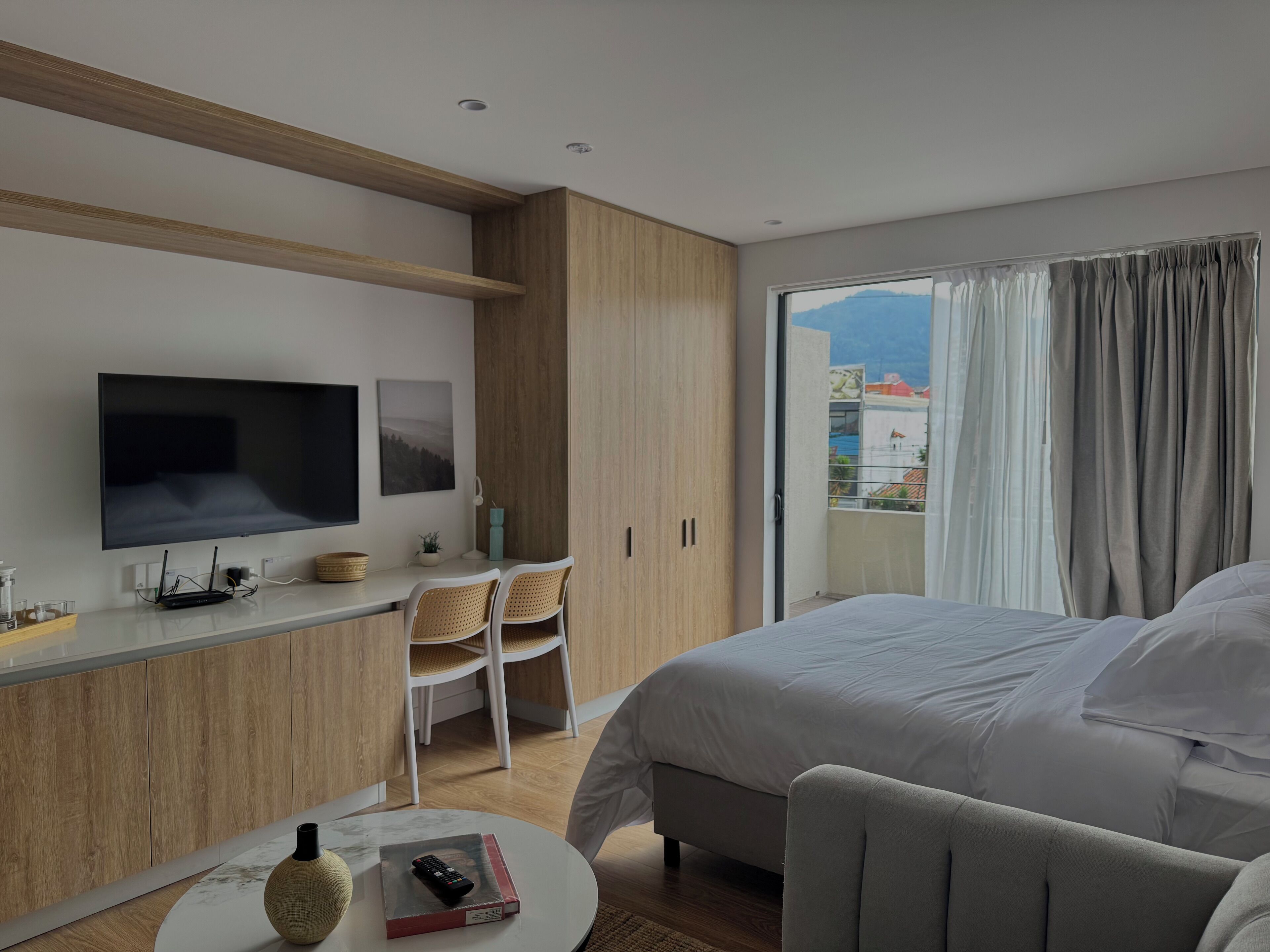 A modern hotel room with a large bed, a TV mounted on the wall, a desk with chairs, and a balcony with curtains.