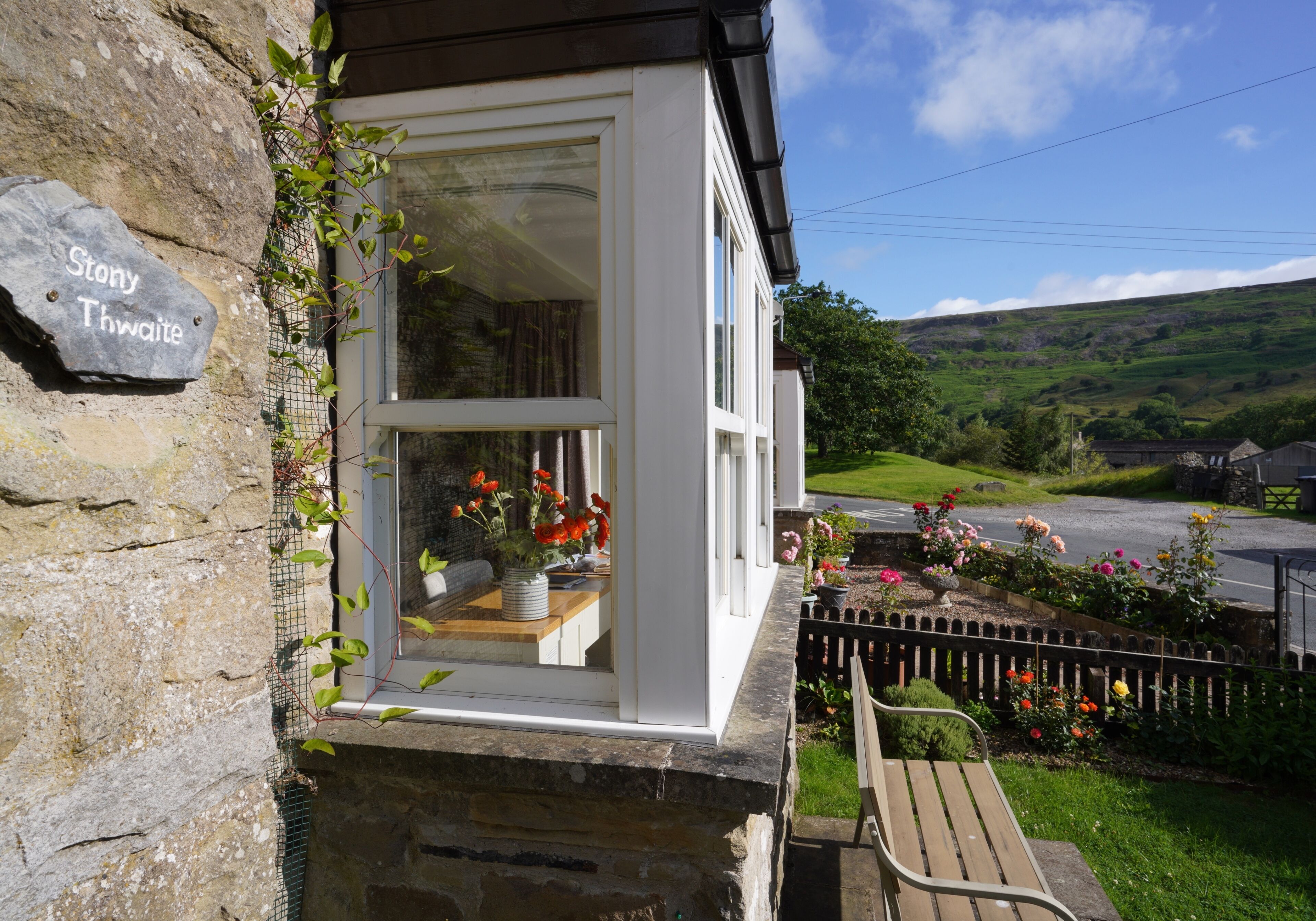 A stone building with a white window, a stone plaque reading 'Stony Thwaite,' and a garden with flowers and a bench.