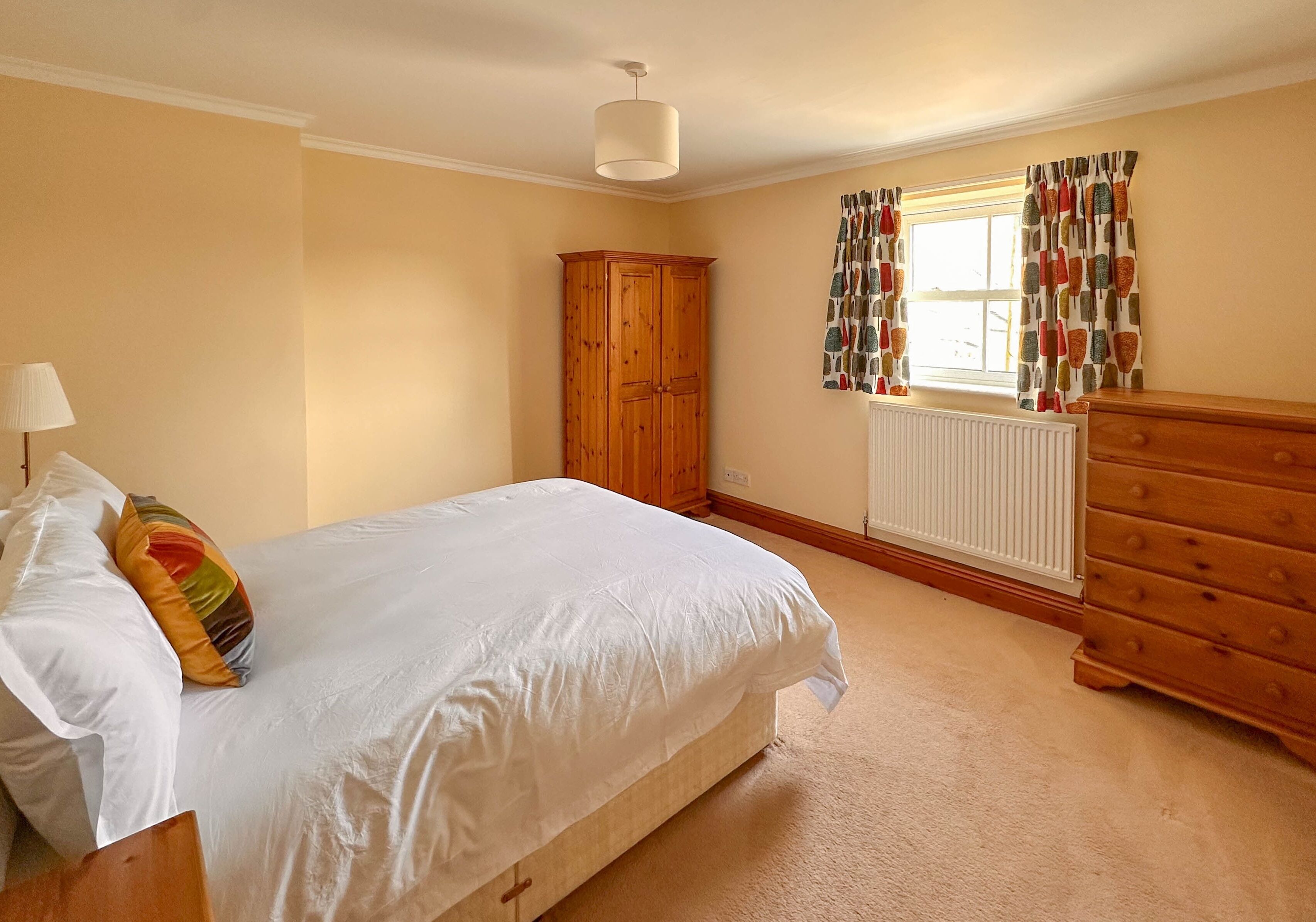 A bedroom with a large bed, a wooden wardrobe, a dresser, a radiator, and a window with patterned curtains.