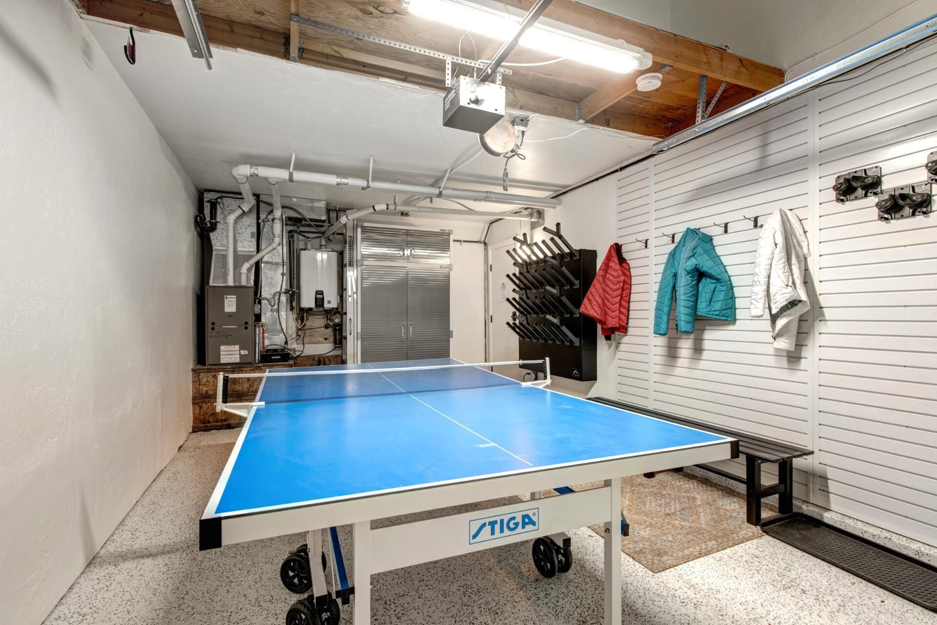 A ping pong table in a room with a white wall and a wooden ceiling. There are jackets hanging on the wall.