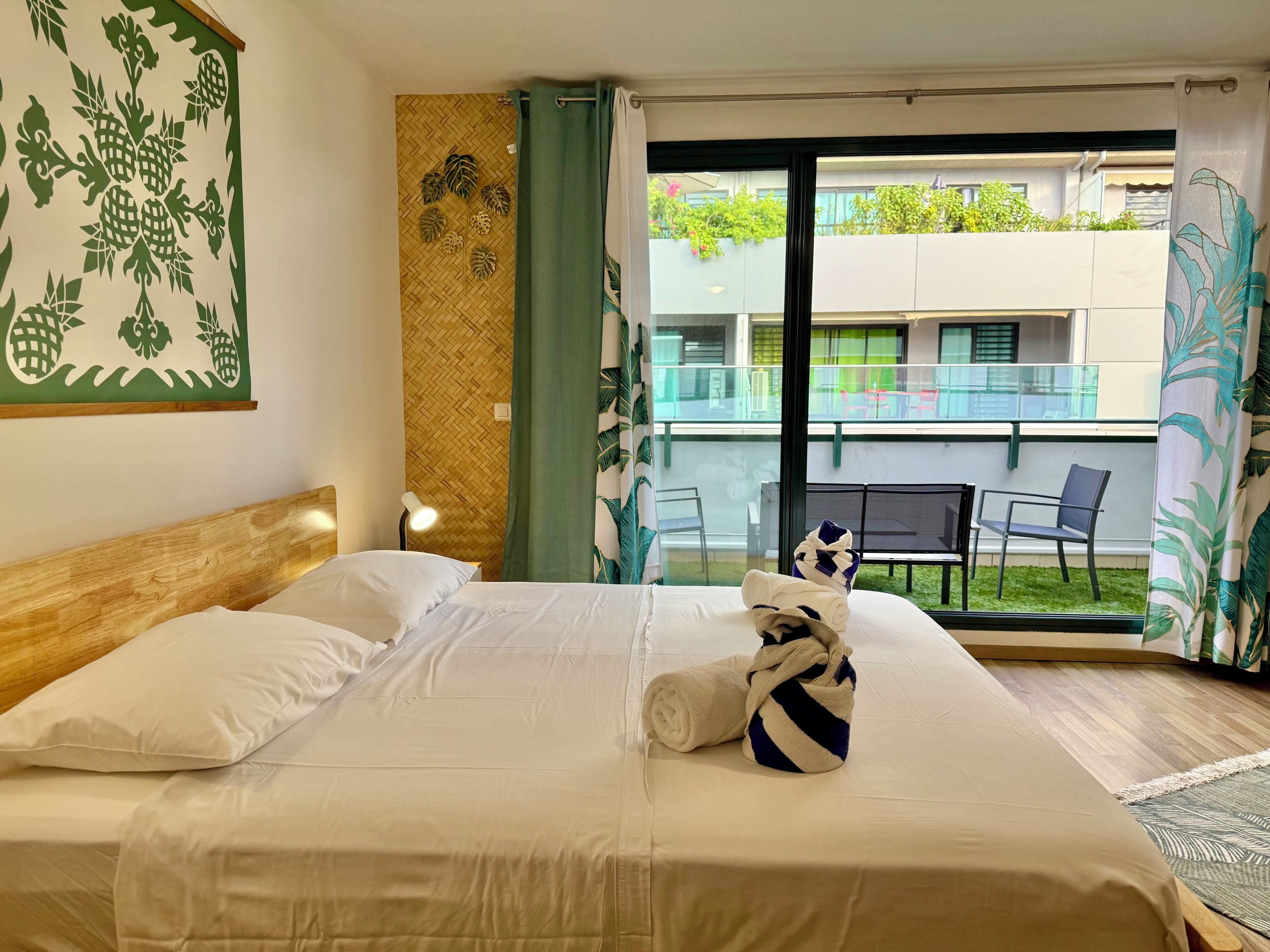 A hotel room with a bed, a nightstand, a lamp, a window with a view of the outside balcony, and a wall with a leaf pattern.
