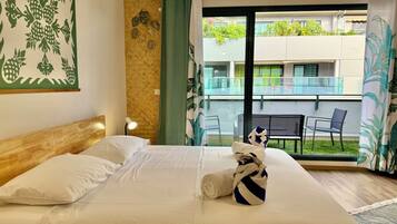 A hotel room with a bed, a nightstand, a lamp, a window with a view of the outside balcony, and a wall with a leaf pattern.