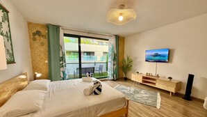 Une chambre moderne avec un lit en bois, un téléviseur fixé au mur et un balcon offrant une vue sur l’immeuble.