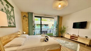 Un lit bien fait avec une tête de lit en bois, une lampe de chevet, une petite plante en pot, une commode en bois sur laquelle se trouve un téléviseur, et une vue sur le balcon extérieur.