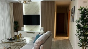 Un salon moderne avec un canapé, une table basse et un téléviseur. On y trouve également une plante en pot et un couloir qui mène à une autre pièce.
