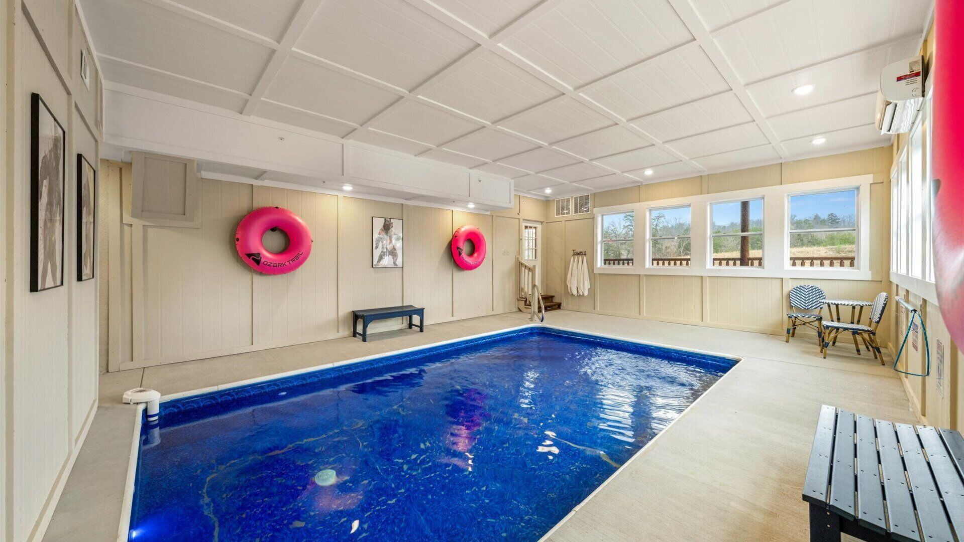 An indoor swimming pool with a bench and two pink inflatable rings, wooden walls, and a view of the outdoors through windows.