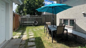 A backyard with a patio table and chairs, a blue umbrella, and a wooden fence. - Cozy & Modern 3BR Lower Unit I Private Entrance, Laundry, Fridge Microwave (Calgary)