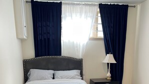 A neatly made bed with a dark headboard, white bedding, and a folded towel on the footboard. A bedside table with a lamp and a dark-colored drawer is visible. - Cozy & Modern 3BR Lower Unit I Private Entrance, Laundry, Fridge Microwave (Calgary)