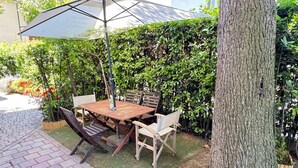 Eine Terrasse im Freien mit einem Holztisch und Stühlen, einem Metallschirm und einer Hecke im Hintergrund.