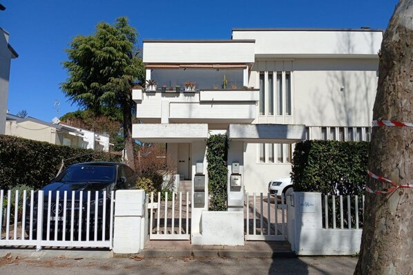 A modern two-story building with a gated entrance, a car parked in front, and a tree to the left.