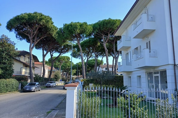 A residential street with parked cars, a white building with balconies, and trees lining the road.