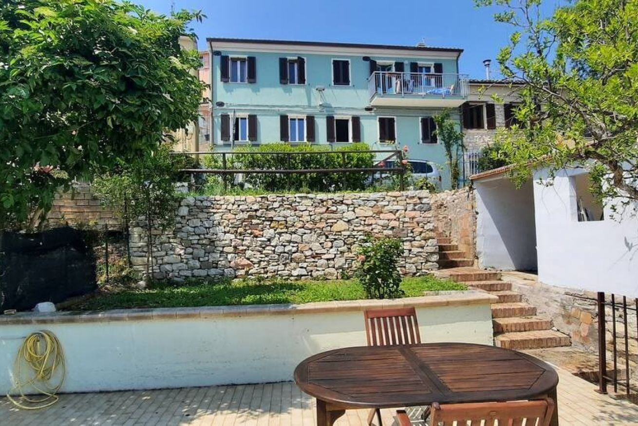 A building with a stone wall, a balcony, and a table with chairs in the foreground.