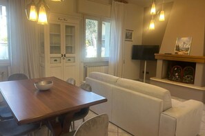 A dining area with a wooden table, white chairs, a white sofa, and a fireplace with a television above it.