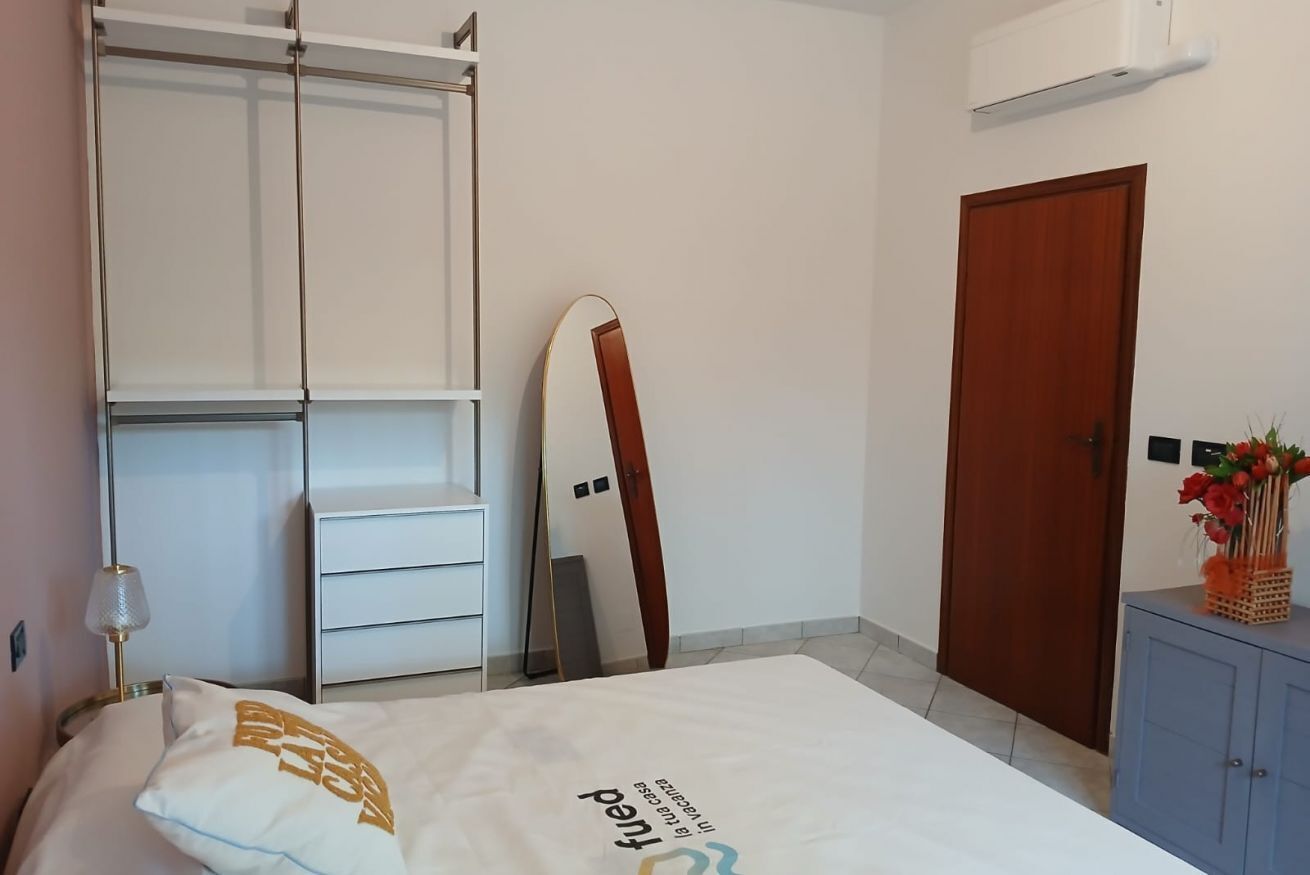 Een slaapkamer met een bed, een kledingkast, een spiegel, een deur en een klein kastje met bloemen.