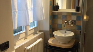 Een badkamer met een wit wastafelmeubel, een raam met witte gordijnen, een betegelde spatwand en een spiegel boven het wastafelmeubel.