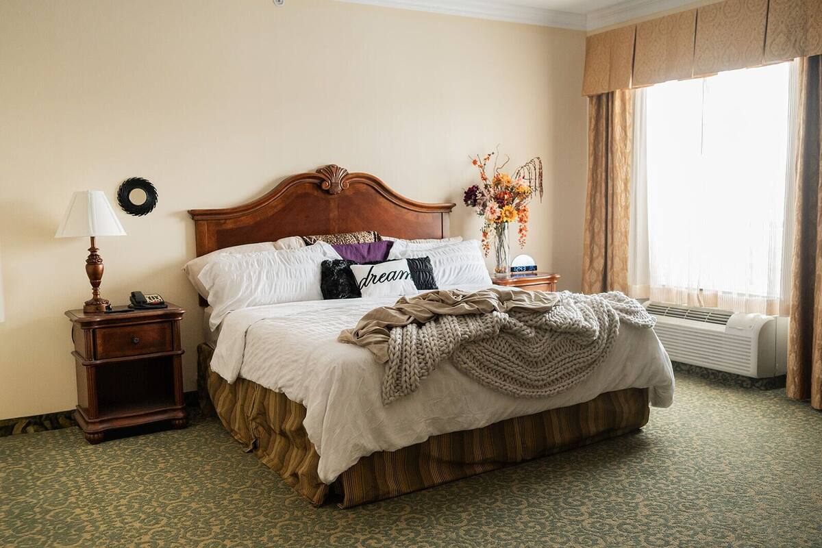 Una habitación de hotel con una cama grande, una mesita de noche con lámpara, un adorno en la pared y una ventana con cortinas.