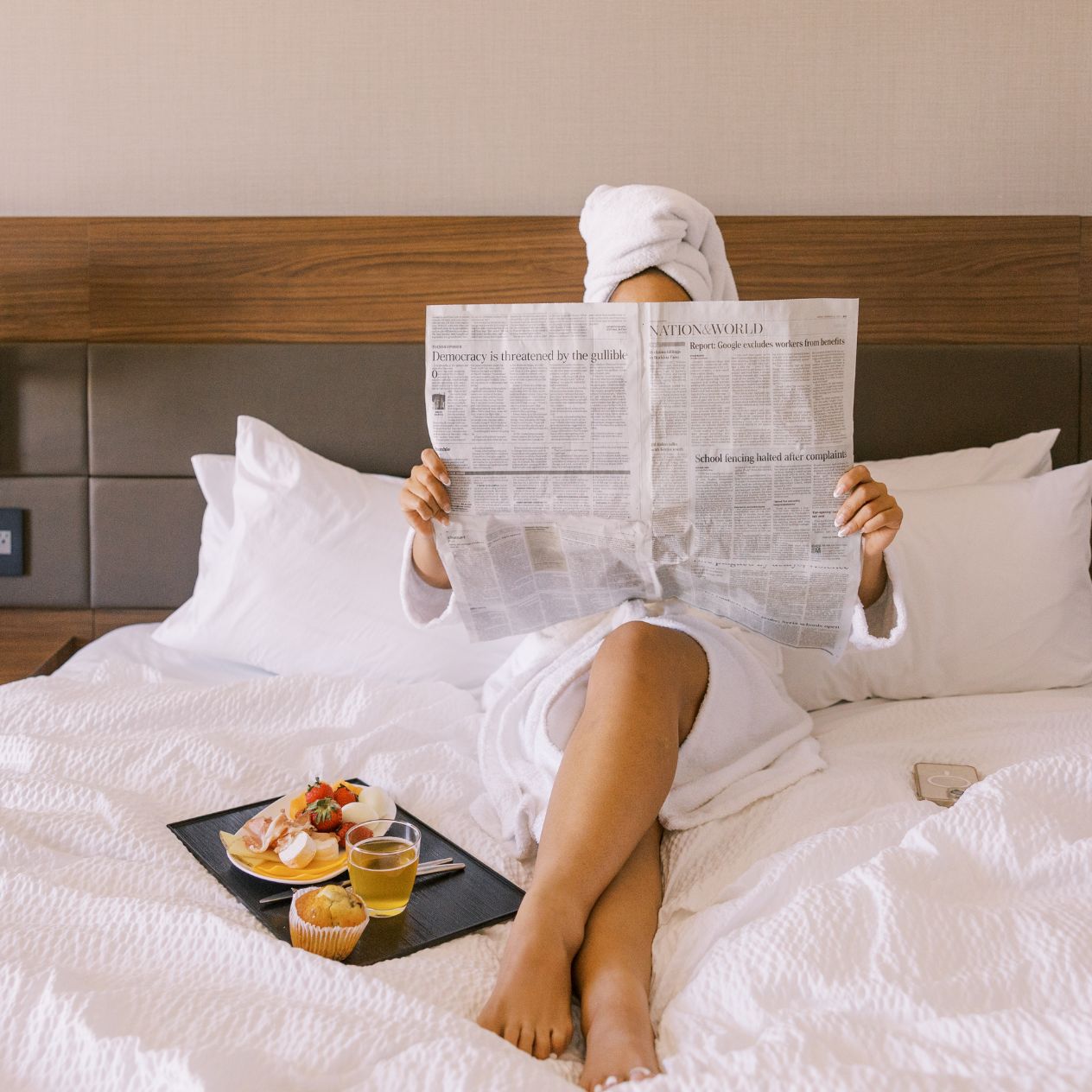 Una persona sdraiata su un letto con un giornale, un piatto di cibo e un bicchiere di bevanda.