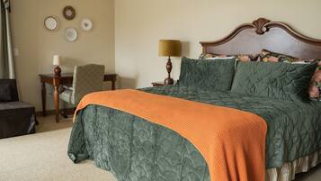 Una cama bien tendida con un edredón verde y una manta naranja, una mesita de noche de madera con una lámpara, y una silla en una habitación con paredes color beige y platos decorativos.