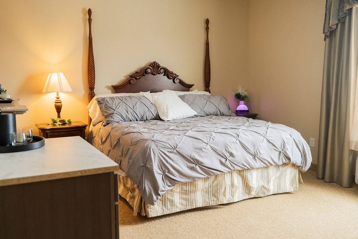 Una cama con dosel, un edredón gris acolchado, una mesita de noche con lámpara y una planta pequeña.