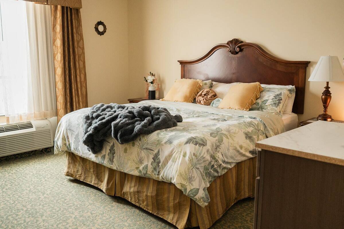 Habitación de hotel con una cama grande, cabecero de madera, mesita de noche con lámpara y una ventana con cortinas.