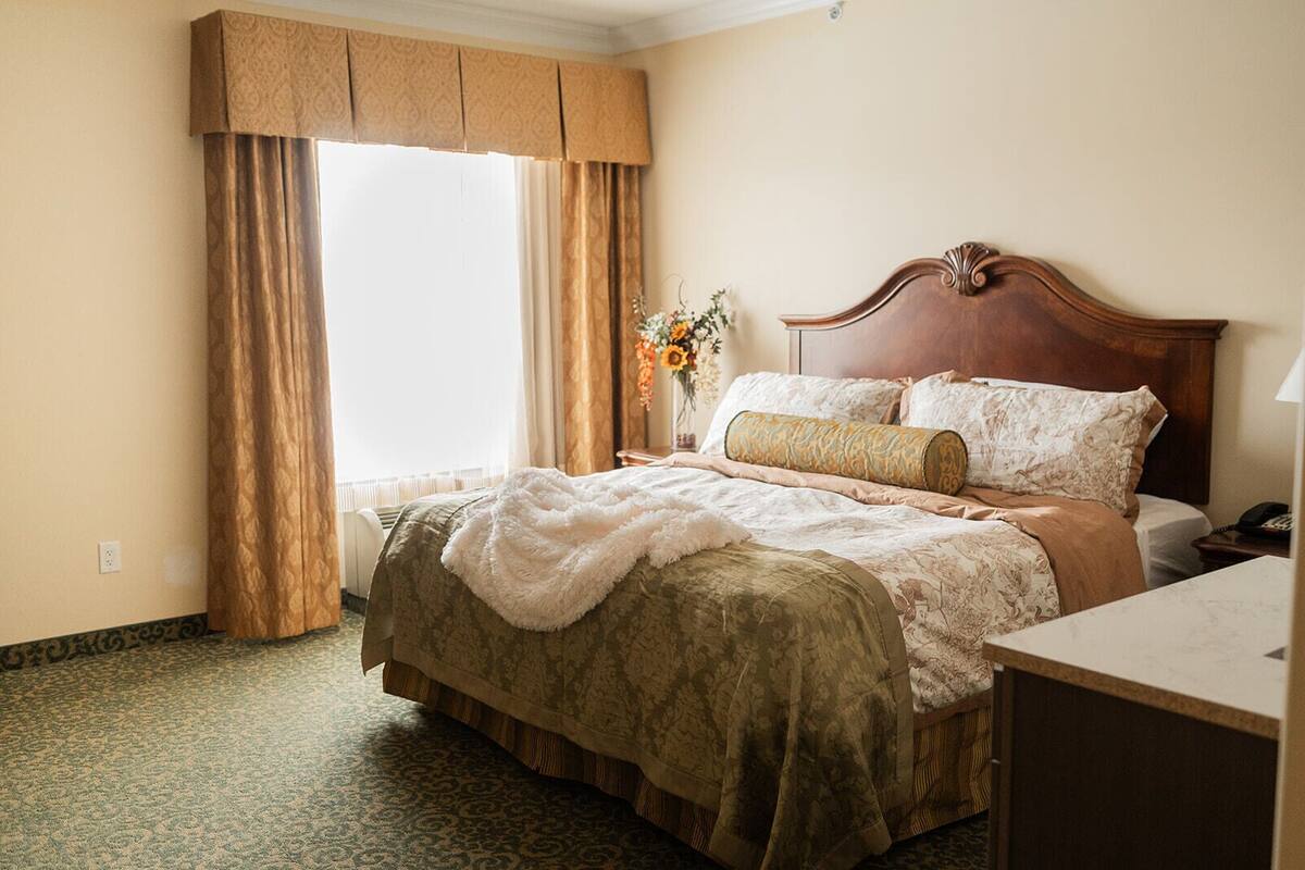 Una habitación de hotel con una cama grande, cabecera de madera, mesita de noche con lámpara y un jarrón con flores.