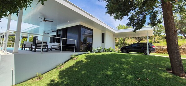 Ein modernes Haus mit überdachter Terrasse, einem Swimmingpool und einem geparkten Auto davor.