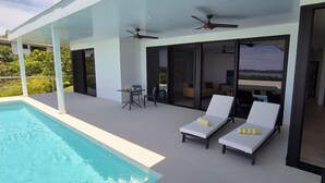 Un área de piscina moderna al aire libre con dos sillones de descanso, una mesa y ventiladores de techo.