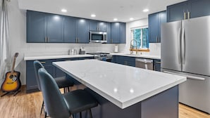 Una cocina moderna con armarios azul oscuro, una isla grande con taburetes altos y electrodomésticos de acero inoxidable, además de vistas al exterior a través de una ventana.
