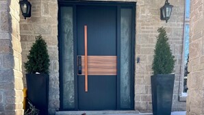 A modern entrance with a stone facade, a dark door with a vertical handle, and two potted plants on either side. - Spacious Retreat in the Pines • Walk to Georgian Bay Beaches (Tiny)