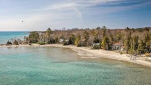 A coastal area with clear turquoise water, a sandy beach, and a forested hill in the background. - Spacious Retreat in the Pines • Walk to Georgian Bay Beaches (Tiny)